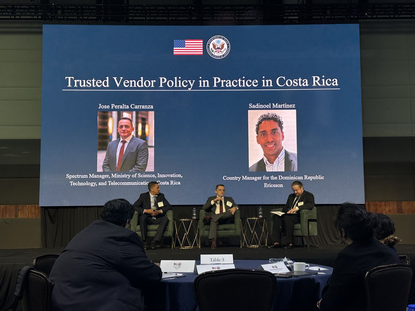 Three panellists on stage in front of a slide introducing the Costa Rica trusted vendor policy