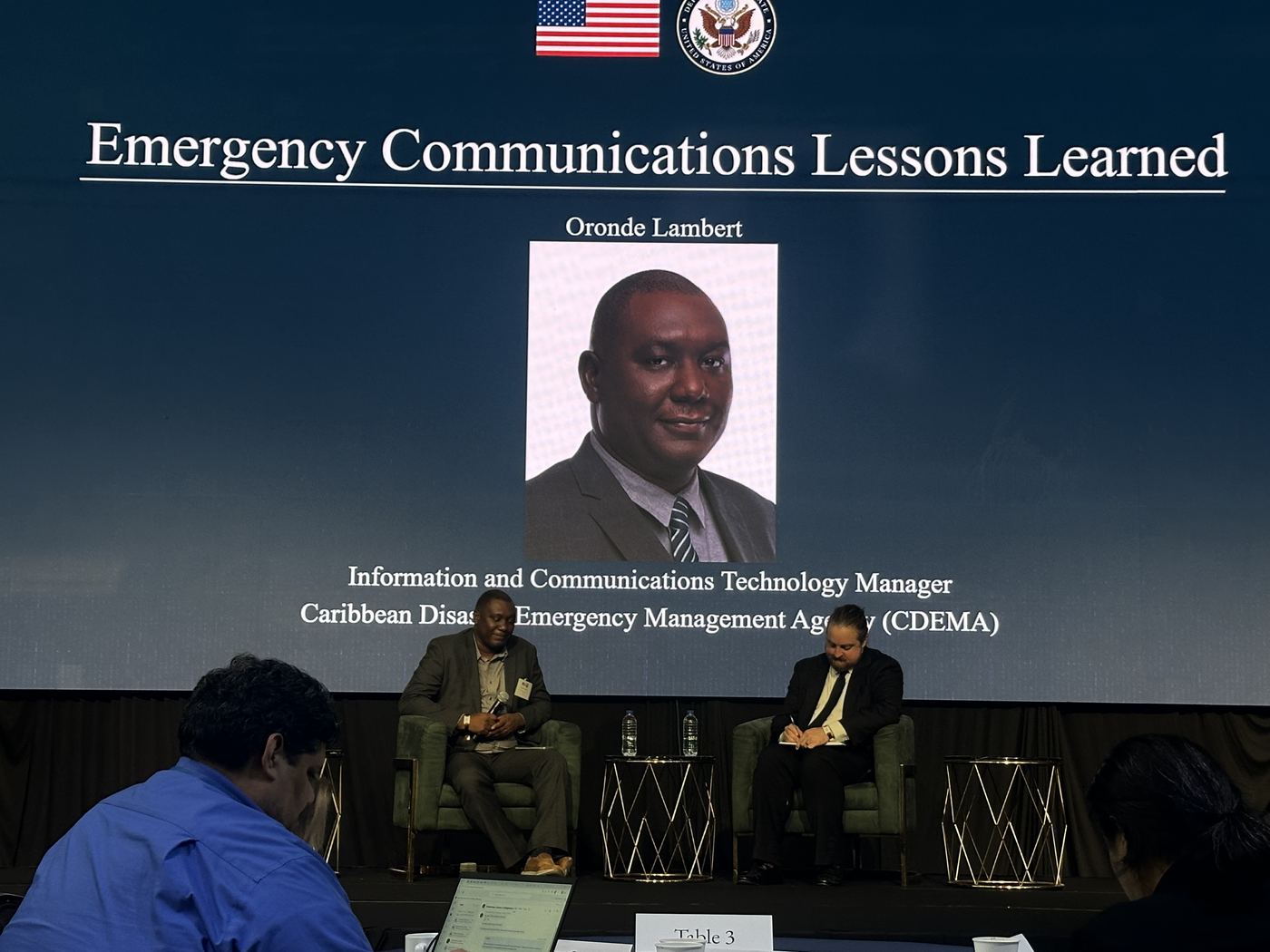 Two speakers on stage in front of a slide introducing Oronde Lambert of CDEMA